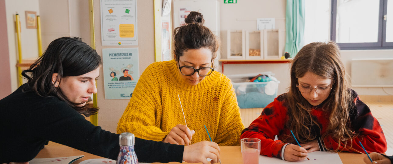 Animatrice à l'écoute d'un·e jeune lors d'une colonie de vacances inclusive, incarnant la pédagogie du care. Le groupe participe à une activité artistique et créative.